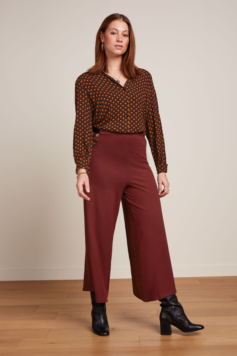 Modell trägt eine weite rostrote Culotte-Hose, kombiniert mit schwarzen Stiefeletten und einer gemusterten Retro-Bluse
