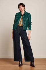 Modell trägt retro-inspirierte Kunstlederjacke in grün, kombiniert mit weiten dunkelblauen Jeans und weinroten Lack-Stiefeletten