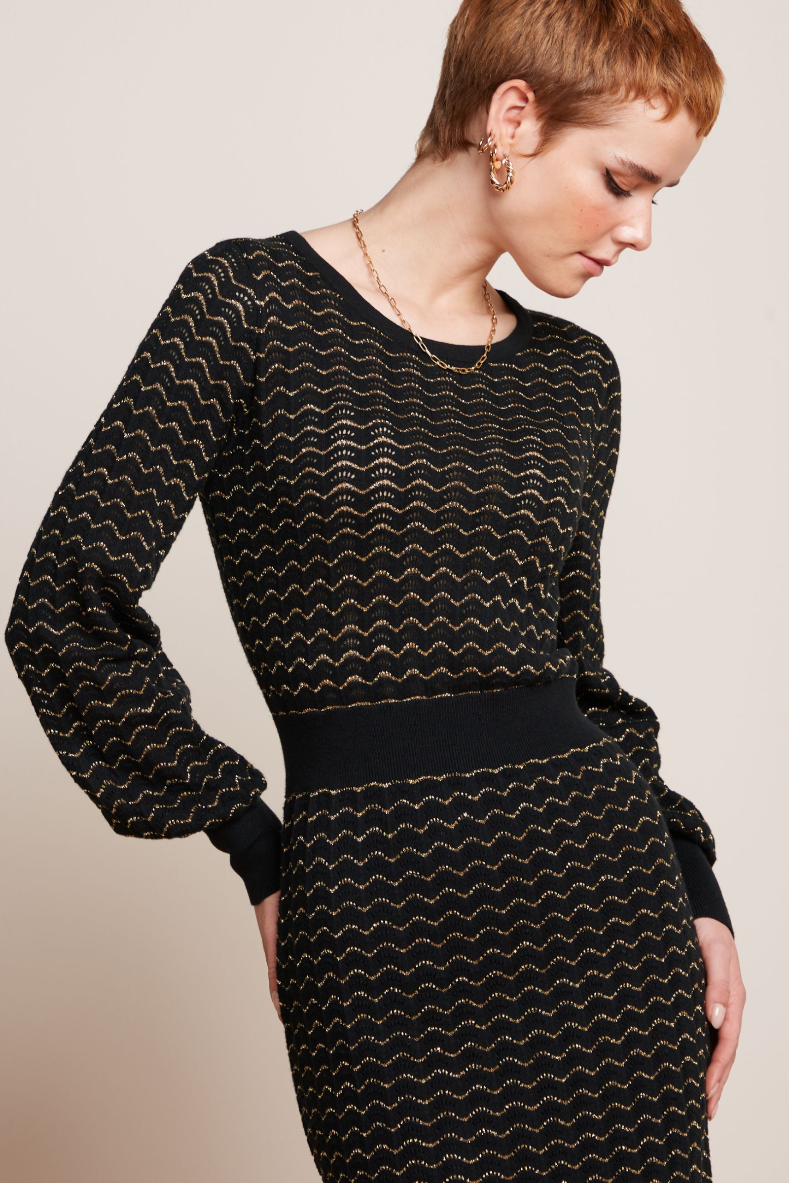 Modell mit Pixie-Frisur trägt elegantes, retro-inspiriertes Kleid mit Glockenärmeln und gold-glitzernden Details,