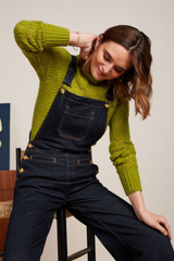 Modell trägt retro-inspirierte denim jeans Latzhose mit goldenen Details, kombiniert mit grünem Lochstrick-Pullover