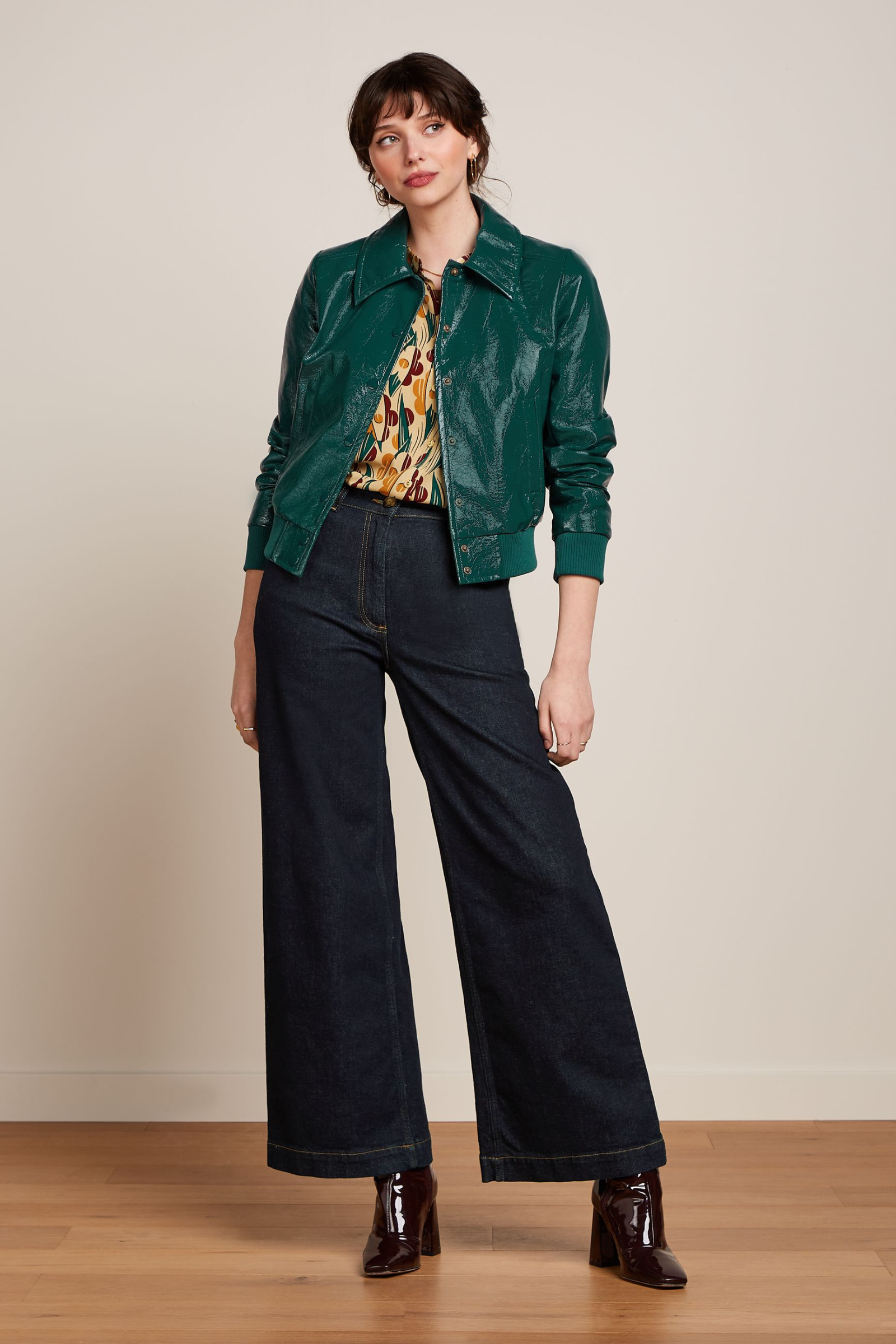 Modell trägt retro-inspirierte Kunstlederjacke in grün, kombiniert mit weiten dunkelblauen Jeans und weinroten Lack-Stiefeletten